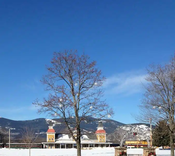 A large building with twin towers and red roofs is set against a snowy landscape with bare trees and mountains in the background, under a clear blue sky.