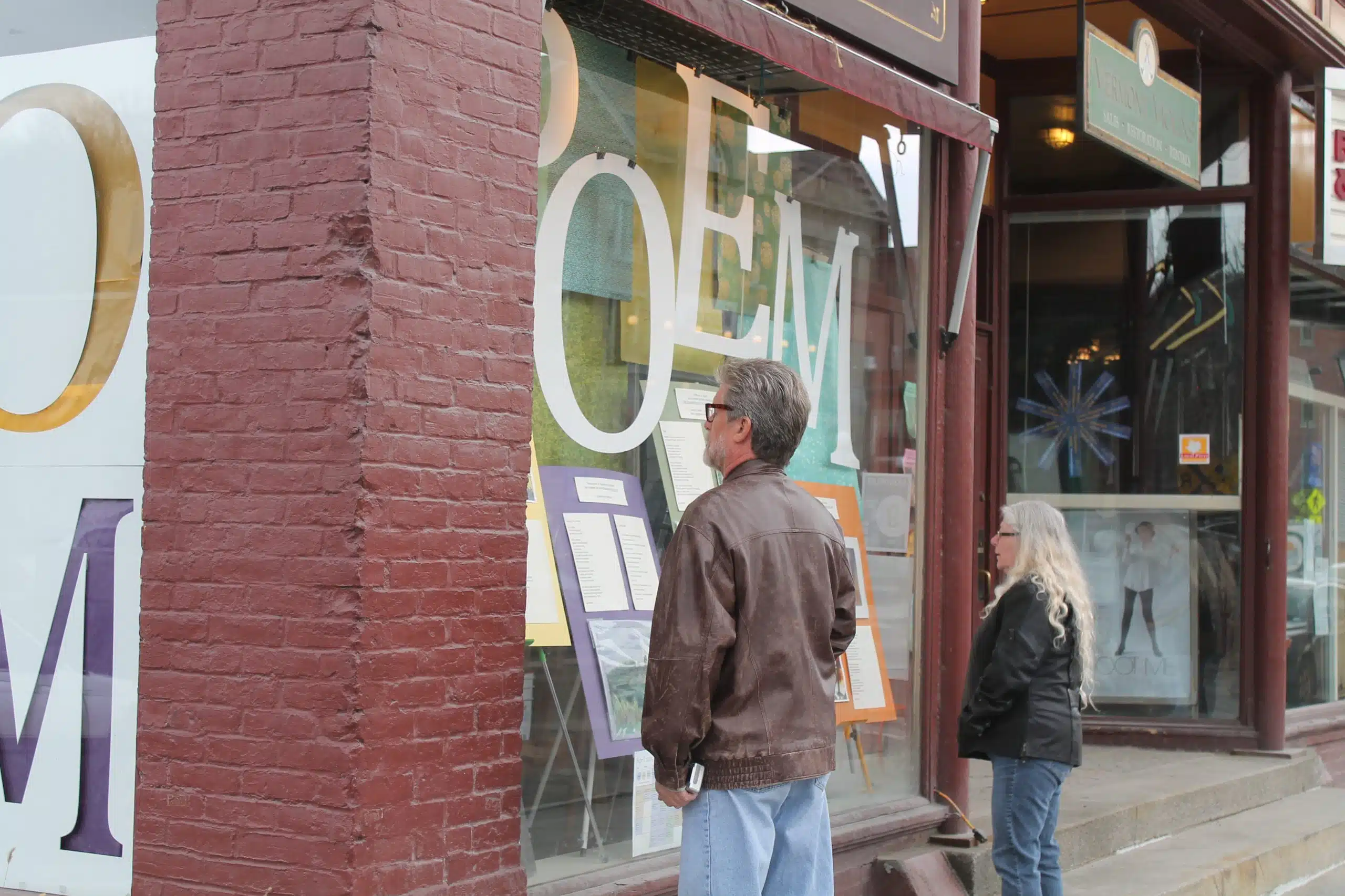 Two individuals stand on a sidewalk, looking at a storefront window display with large "POEM" letters and several posted documents.