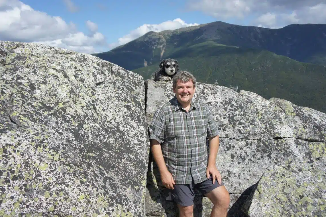 A man in a plaid shirt and shorts stands on a rocky mountain ledge with a small dog beside him. A mountainous landscape is visible in the background under a partly cloudy sky.