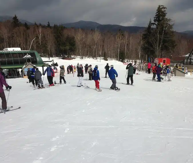 A group of people in winter gear are skiing and snowboarding on a snow-covered slope near a forested area with a ski lift in the background.