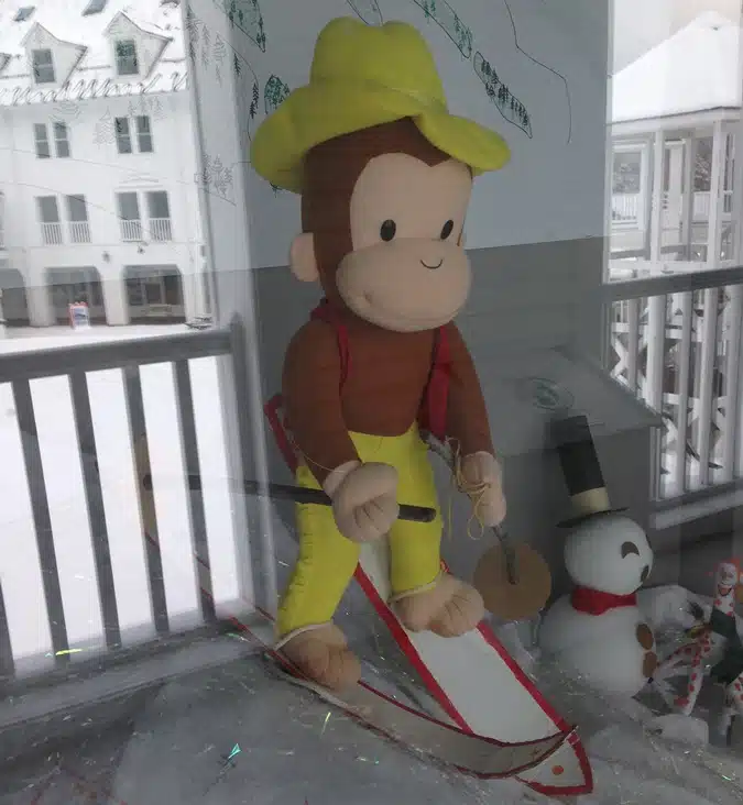 A stuffed monkey toy dressed as a skier with a yellow hat and red suspenders is posed on a ski slope. Snowy scenery and a small snowman are visible in the background.