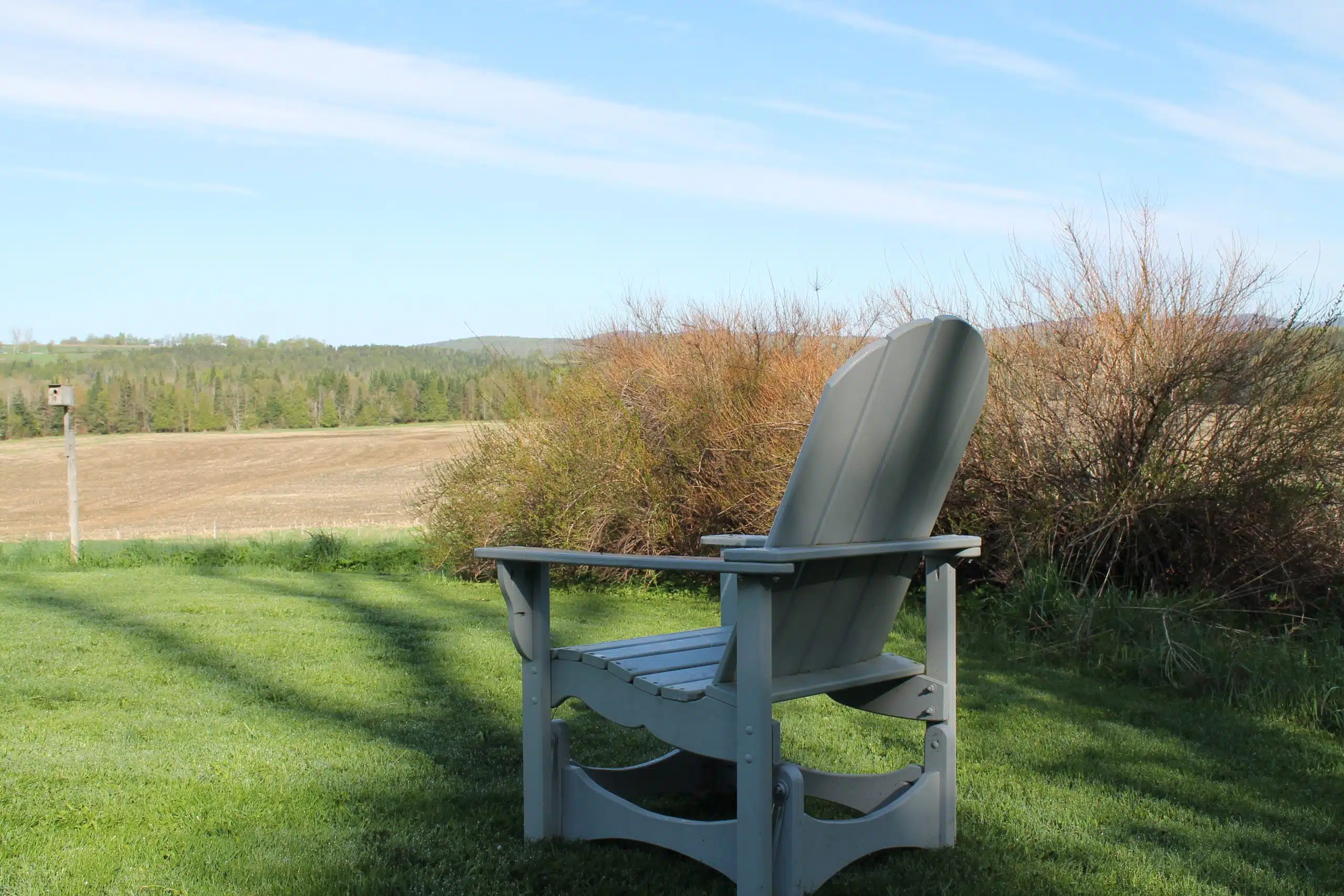 A gray Adirondack chair sits on a grassy lawn, facing a scenic view of open fields and wooded hills under a clear blue sky.
