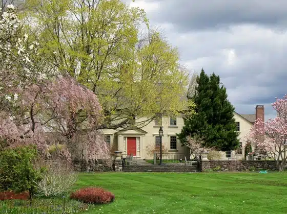 A two-story house with a red door is surrounded by flowering trees and a large green lawn under a cloudy sky.