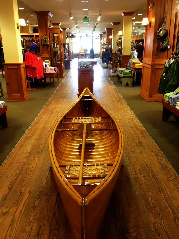 A wooden canoe displayed in the center aisle of a retail store, surrounded by racks of clothing and outdoor gear. The store features wooden furnishings and well-lit interior.