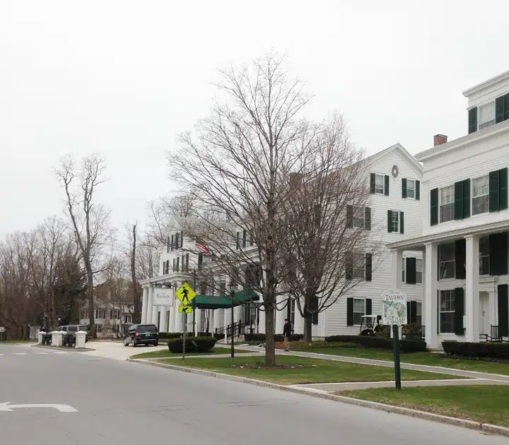 A street scene showing white buildings with green shutters and trees without leaves. Sidewalks, a pedestrian crossing sign, and a road are in view. The sky is overcast.