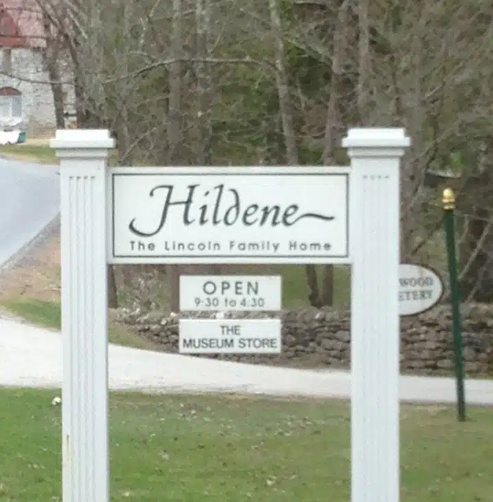Sign for Hildene, the Lincoln family home, indicating it is open from 9:30 to 4:30. Another sign below points to the museum store. Trees and a road are visible in the background.