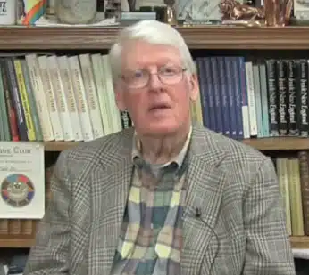 An older man with white hair and glasses is seated in front of a filled bookshelf, wearing a plaid shirt and a checkered blazer.