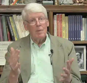 An elderly man with white hair and glasses, wearing a green shirt and beige blazer, sits in front of a bookshelf, gesturing with his hands while speaking.