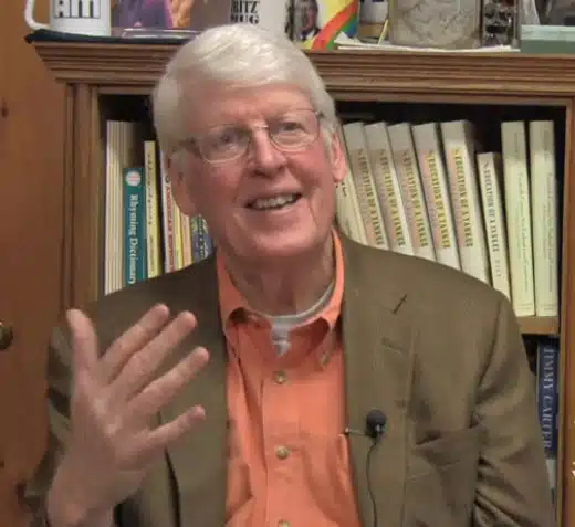 An elderly man with white hair and glasses is gesturing with his right hand while sitting in front of a bookshelf filled with books. He is wearing a brown blazer and an orange shirt.
