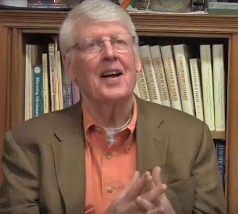 An older man with white hair and glasses is smiling and speaking. He is wearing a brown blazer and an orange shirt. Behind him is a bookcase filled with various books.