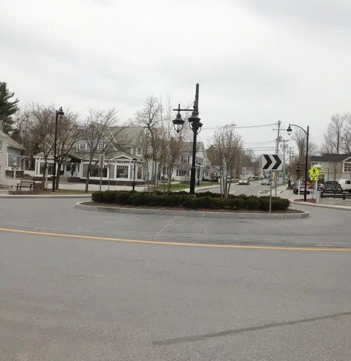 A small town roundabout with street lamps, bare trees, surrounding buildings, a pedestrian crossing sign, and overcast skies.