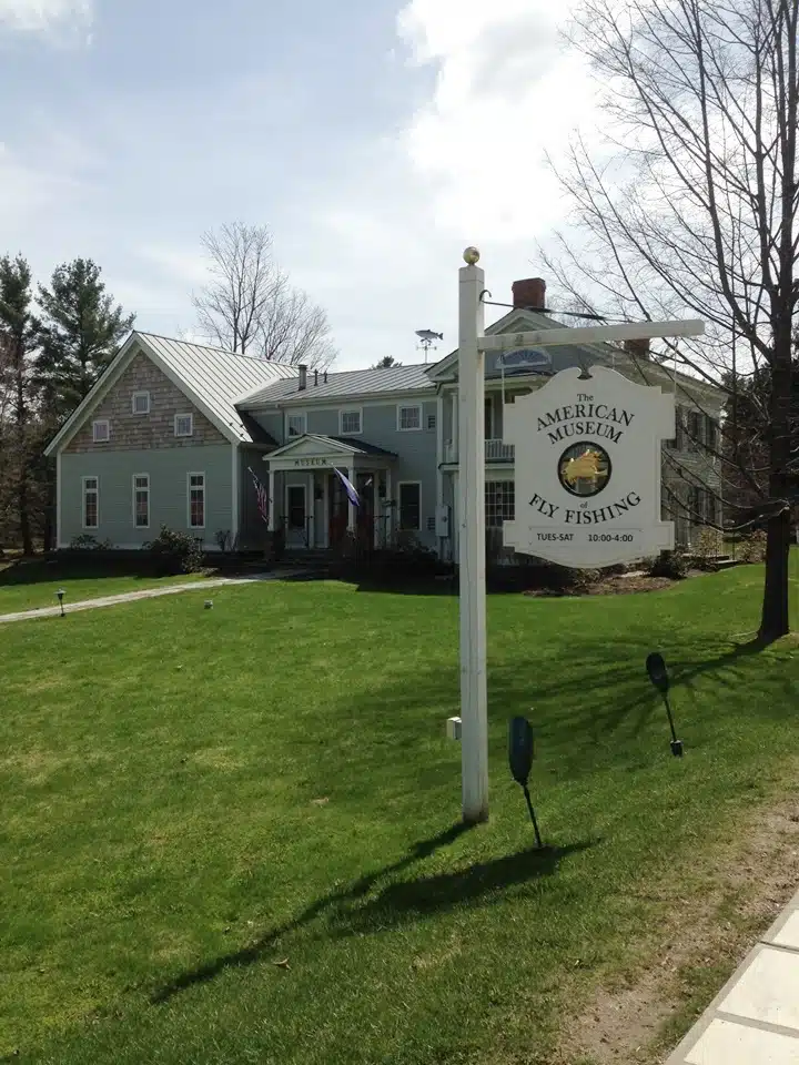 A green building with white trim houses the American Museum of Fly Fishing. A white sign with information stands on a grassy lawn with trees in the background.
