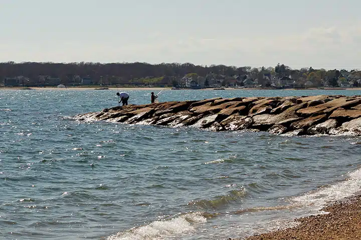 Two people stand on a rocky jetty stretching out into a body of water with a distant shoreline and houses in the background.