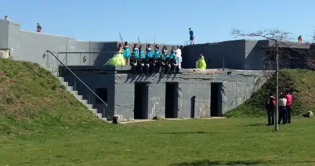 A group of people dressed in bright costumes sit on top of a concrete structure, with others standing nearby on the grass and a clear blue sky above.