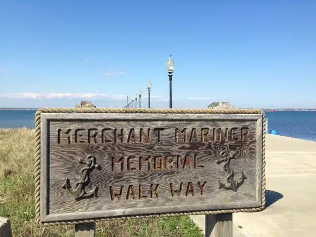 Wooden sign reading "Merchant Mariner Memorial Walk Way" with anchor imagery in front of a oceanfront walkway and lampposts.