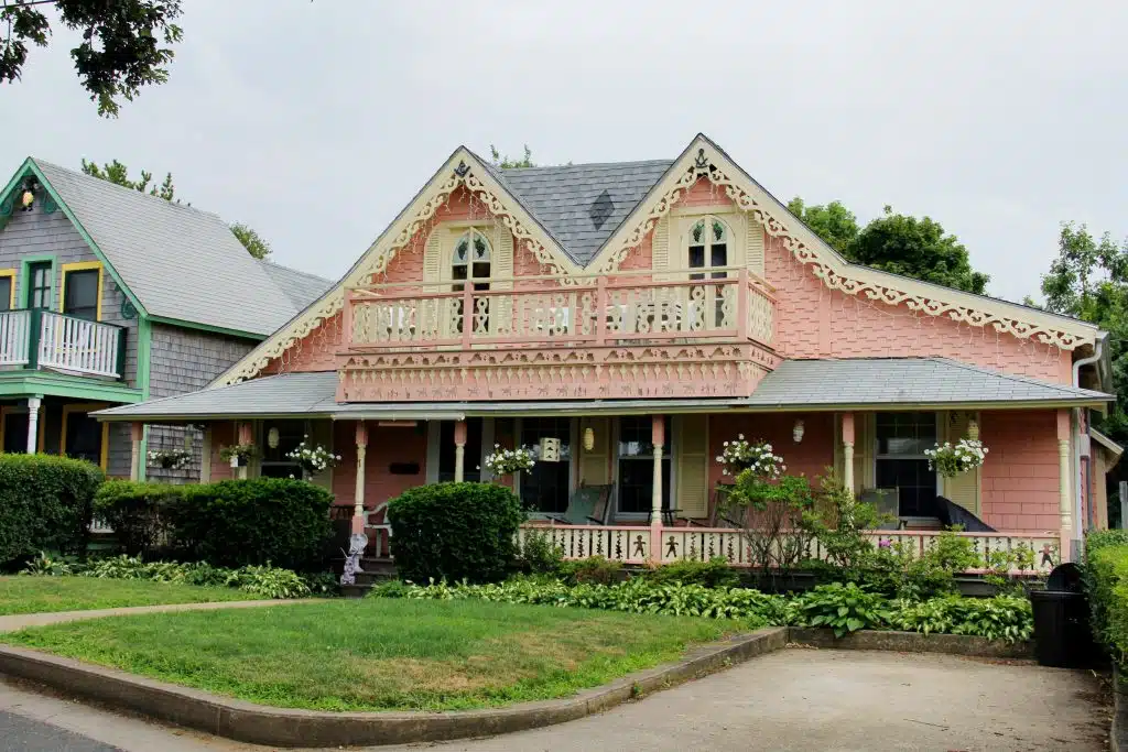 A charming, two-story, pink house with ornate white trim, a front porch, balcony, and well-maintained garden, located beside a gray house with similar decorative features.