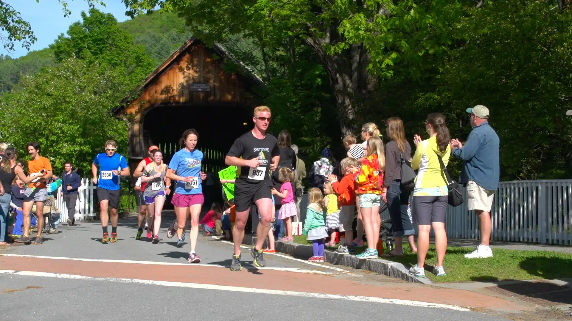 Runners participate in a race near a covered bridge while spectators cheer from the sidelines on a sunny day.
