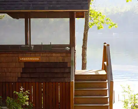 A wooden lakeside cabin named "Ardenwood," featuring stairs leading to an elevated porch, surrounded by trees and near a body of water.
