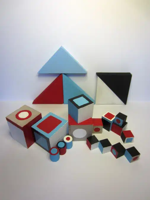 Various geometric wooden blocks in different shapes and colors (blue, red, black, white) are arranged on a white surface against a light background.