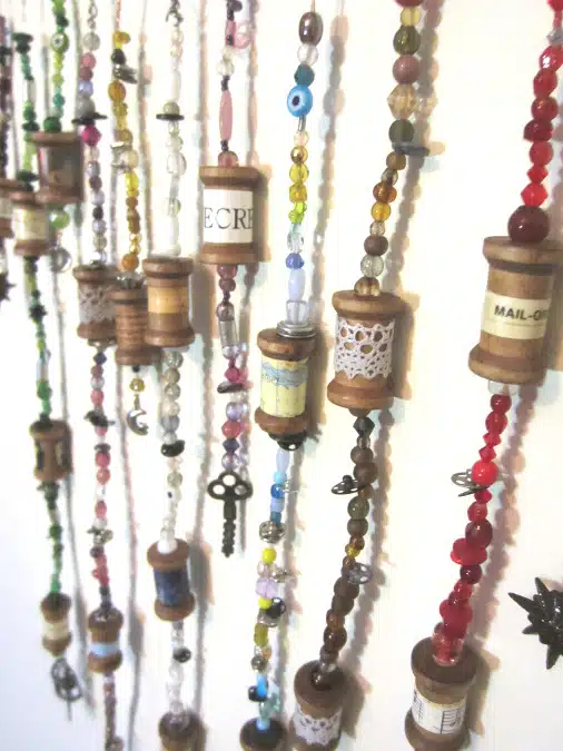 Handcrafted bead and spool strings hang vertically against a white wall, featuring various colored beads and repurposed wooden spools.