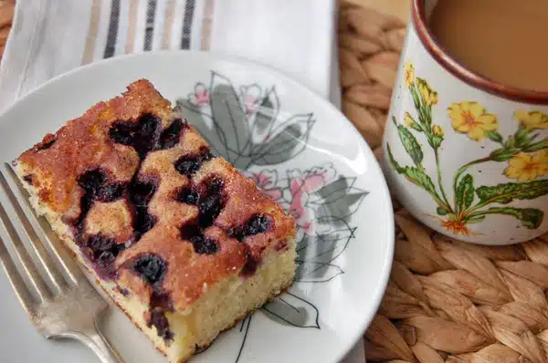 A slice of blueberry cake on a floral plate with a fork, next to a mug of coffee on a woven placemat. Blueberry recipes