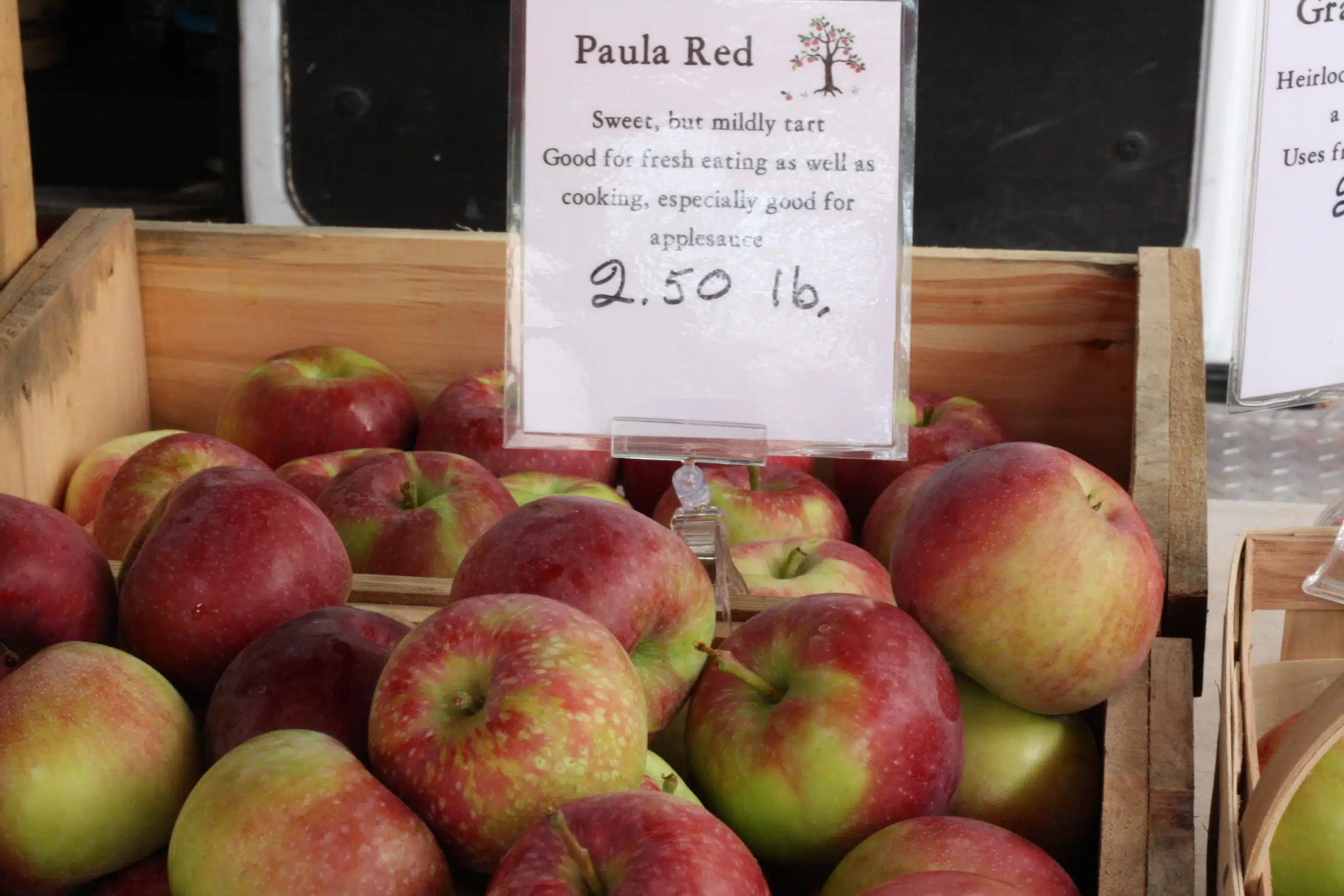 Apples labeled "Paula Red" on display in wooden crates at a market with a sign indicating they are priced at $2.50 per pound and noting their suitability for fresh eating and cooking, especially for applesauce.