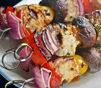 Two grilled skewers with chunks of meat, red onions, bell peppers, mushrooms, and yellow tomatoes served on a white plate.