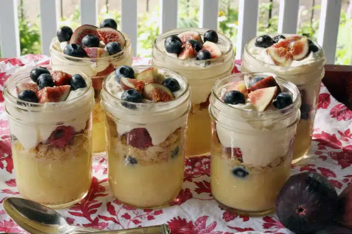 Six glass jars filled with layered dessert, topped with fresh figs and blueberries, are arranged on a red and white tablecloth with a silver spoon and a fig beside them.