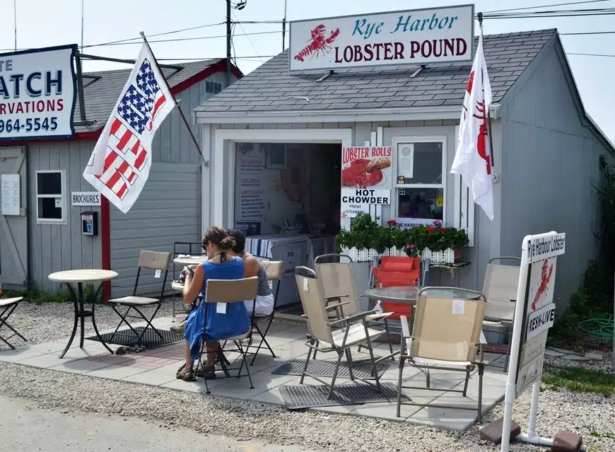 A small, gray, seaside lobster shack with an outdoor seating area featuring tables and chairs. Two people are seated at a table. Flags and signs indicate the sale of lobster rolls and hot chowder.