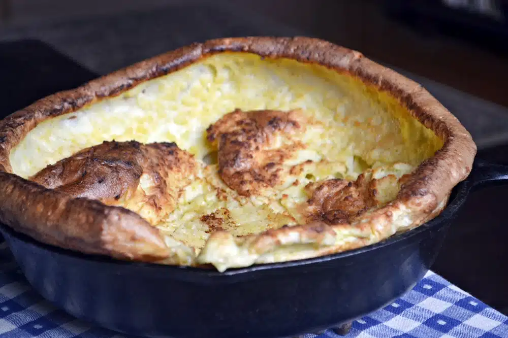 A Dutch baby pancake in a cast iron skillet, with a golden-brown, slightly puffed and crispy edge, placed on a blue and white checkered tablecloth.