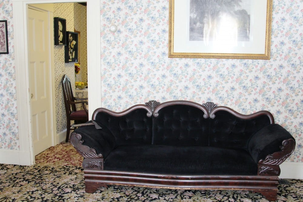 A black vintage sofa in a room with floral wallpaper and a framed painting. An adjacent room with a dining table and chairs is visible through the open door.