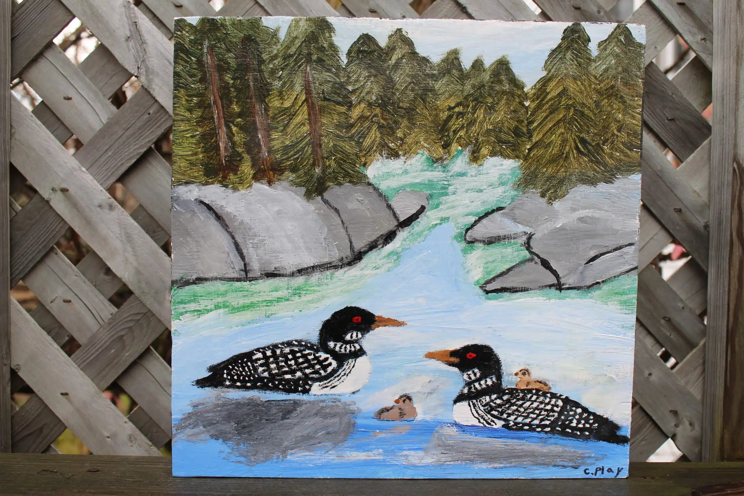 A painting depicts two loons and their chicks on a serene lake surrounded by trees and rocks.