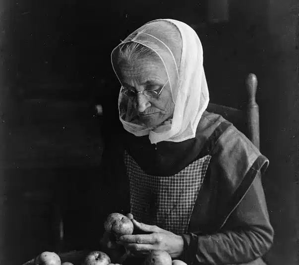 An elderly woman wearing a headscarf and glasses sits at a table sorting apples.