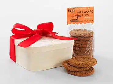 A box with a red ribbon next to a pack of molasses clove cookies and some cookies placed in front.
