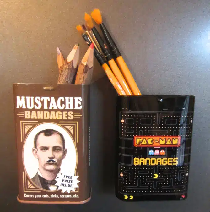 Two containers hold pencils and paintbrushes. One tin is labeled "Mustache Bandages" and the other "Pac-Man Bandages.