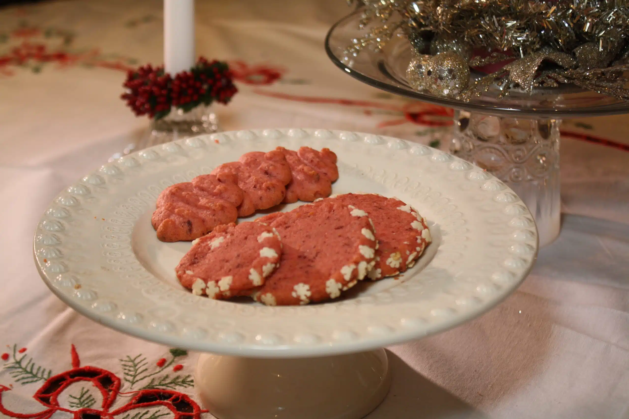 A white plate containing four pink cookies, two shaped as mittens and two round ones with white decorations around the edges, displayed on a white tablecloth with a candle and tinsel in the background.