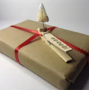A brown paper-wrapped gift with a red ribbon is decorated with a miniature Christmas tree attached to a clothespin.