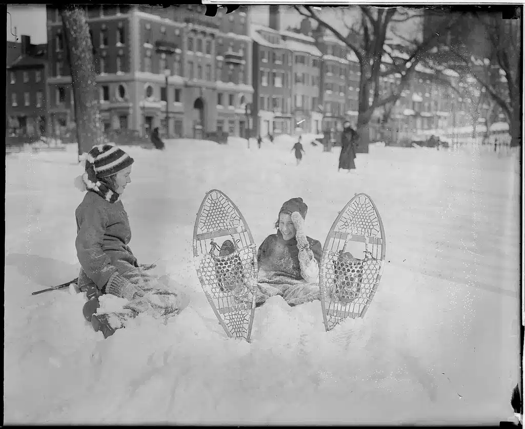 Two individuals sit in the snow with snowshoes on the ground in front of them. Snow-covered buildings and trees are visible in the background, along with other people.