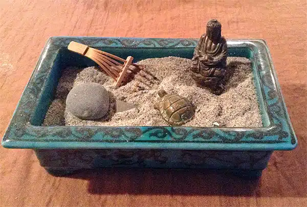 A tabletop Zen garden featuring sand, a rock, a small Buddha statue, a miniature turtle figurine, and a tiny rake in a rectangular, decorated container.