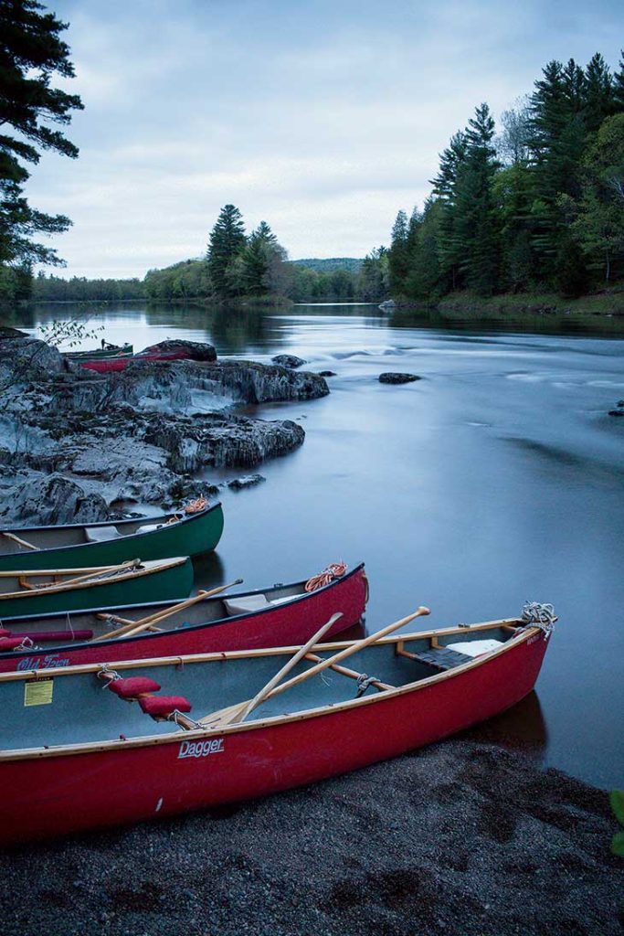 Three canoes are moored on a riverbank, with calm water flowing amidst rocks and a backdrop of dense trees under a cloudy sky.