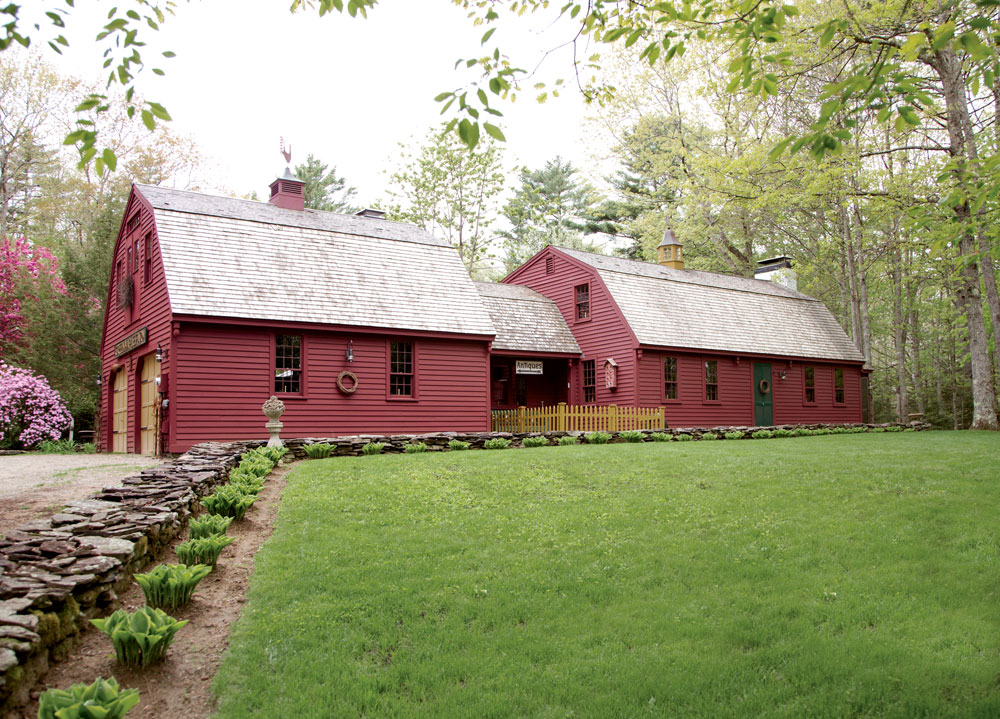 A red Colonial-style house with a gray shingled roof, surrounded by a green lawn, stone walkway, and trees.