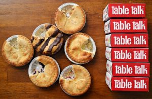 Six small pies, including one chocolate drizzled pastry, are laid out on a table next to a stack of seven red boxes labeled "table talk.