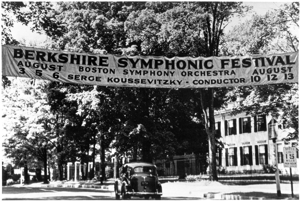 Part of a collection of Tanglewood historic photographs, this images shows a banner hung across a tree-lined street announces the Berkshire Symphonic Festival with the Boston Symphony Orchestra, conducted by Serge Koussevitzky, on August 3-6 and 10-13. A car is parked below.