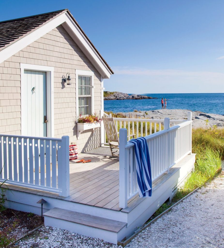 A small beige beach cottage with a white porch overlooks the ocean, with a towel draped over the railing and two people in the distance.