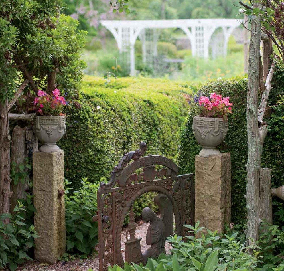 Stone Acres’ side gate is flanked by colorful begonias in urns, as rambling roses climb the arbor overhead; the path leads visitors through neatly trimmed 170-year-old boxwood hedges into a traditional English garden.