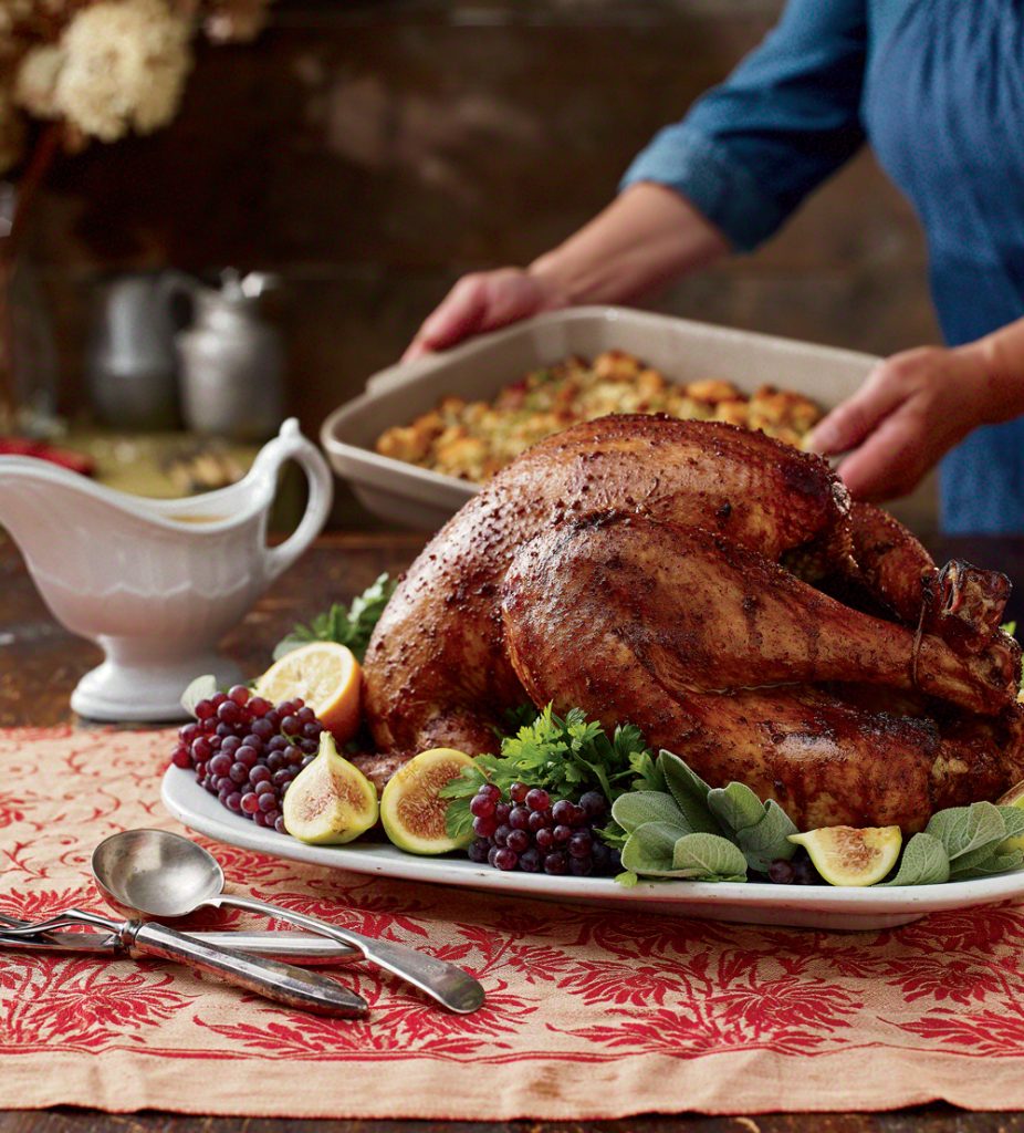 Roasted turkey on a platter garnished with figs and grapes, with a side dish in the background.