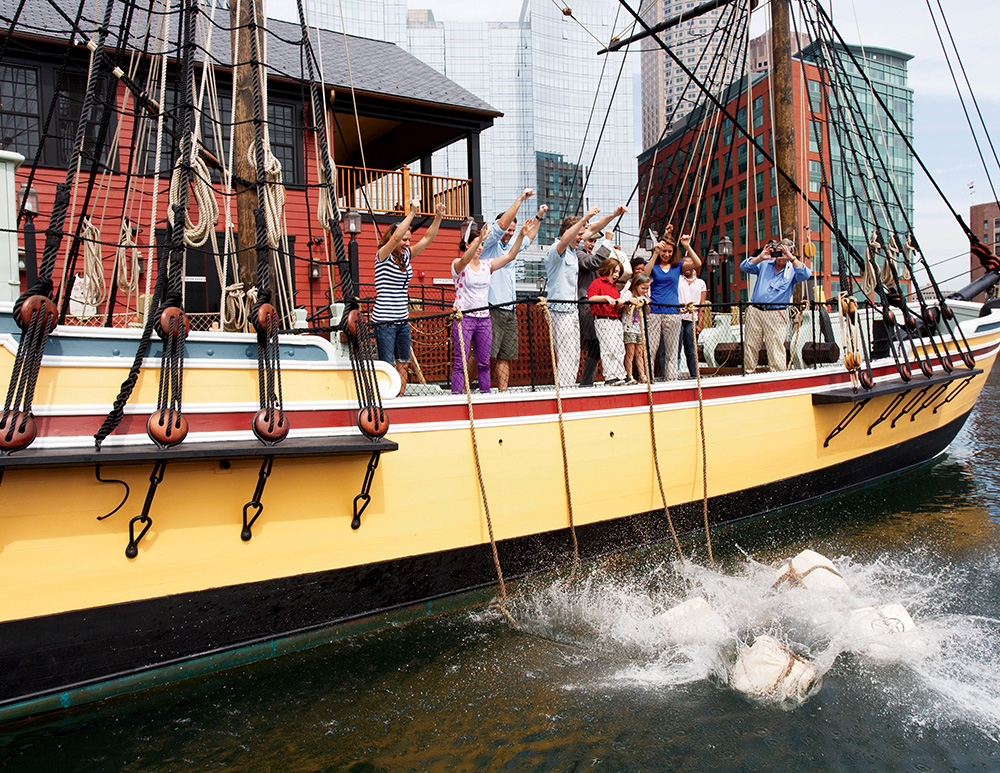 People reenacting the Boston Tea Party by throwing crates off a historic ship at a dock with buildings in the background.