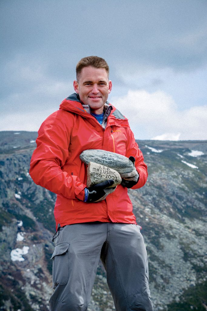 A man in a red jacket stands outdoors on a mountain, holding a rock with inscriptions.