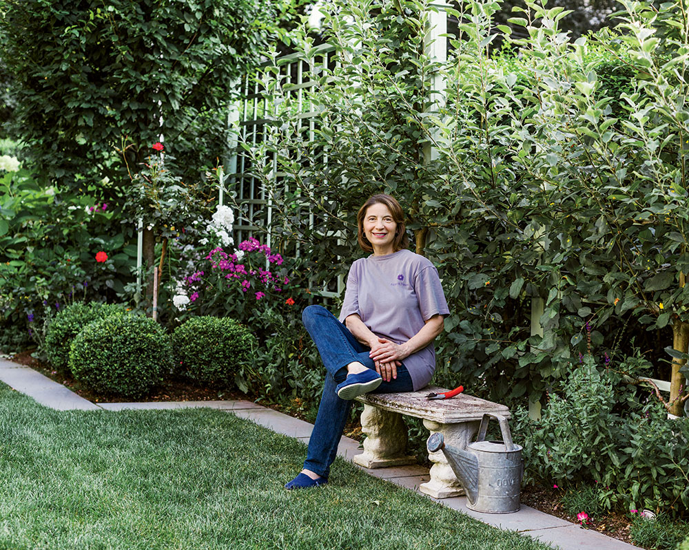 Person seated on a bench in a garden, surrounded by lush green foliage and flowers. A watering can and gardening tool are placed nearby on the ground.