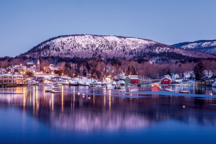 In Midcoast Maine, Camden Harbor’s waters reflect the glimmer of early-evening lights, with the snowy expanse of Mount Battie in the background.